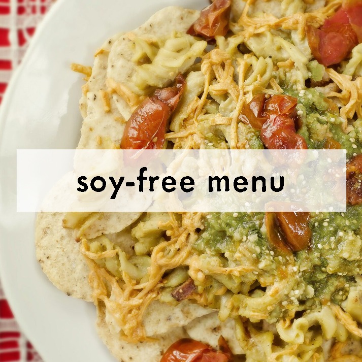 A plate of pasta topped with cherry tomatoes, green pesto, and cheese is labeled "soy-free menu" on a white overlay, placed on a red-patterned cloth.