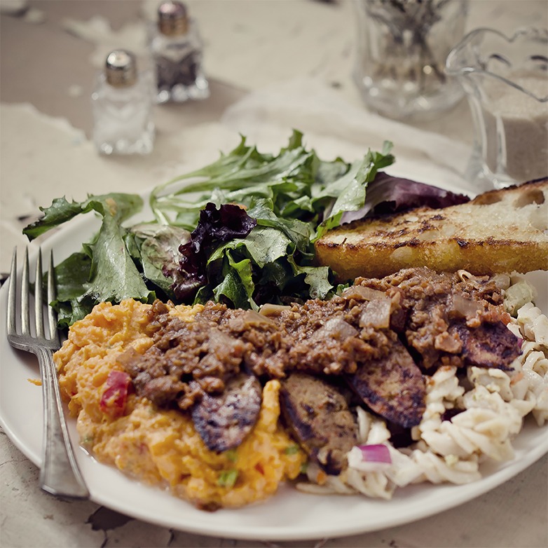 A plate of colorful food featuring meat topped with brown sauce on mashed potatoes, pasta salad, green salad, and grilled bread, conveying a rustic, hearty meal.